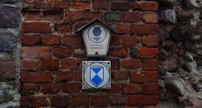 Man sieht zwei weiße Schilder auf einer Wand aus roten Ziegelsteinen. Auf dem oberen Schild steht: Geschütztes Bodendenkmal. Auf dem unteren Schild steht: Denkmal.