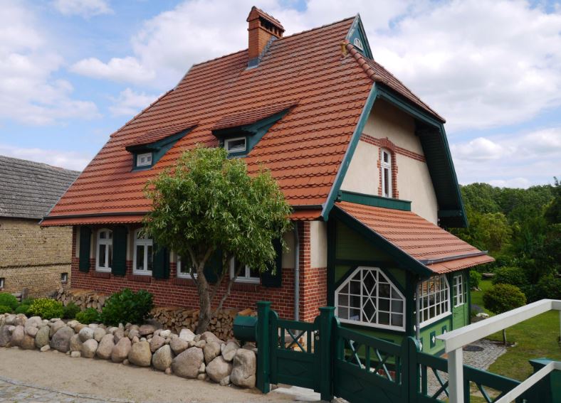 Ein altes Haus. Das Haus ist sehr schön. Es hat ein rotes Dach aus Ziegeln. Vor dem Haus steht ein kleiner grüner Baum. Und ein Zaun aus runden Steinen.