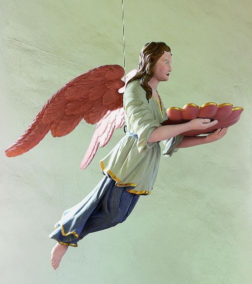 Eine Engels-Figur aus Holz. Der Engel hat rote Flügel und lange braune Haare. Er hängt von der Decke einer Kirche. Er hält mit seinen Händen eine Schale. In der Schale ist Wasser. Mit dem Wasser wurden Menschen getauft.