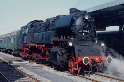 Ein alter Zug. Der Zug ist schwarz und rot. Neben dem Zug ist eine Dampfwolke. Früher wurden die Räder durch Dampf bewegt. So konnte der Zug fahren. Ein anderes Wort dafür ist: Dampf-Lokomotive.