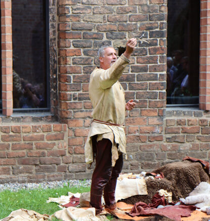 Ein Mann in einer Tunika und Lederhose hält ein Werkzeug, mit dem in der Steinzeit Kleidung aus Tiersehnen gefertigt wurde.